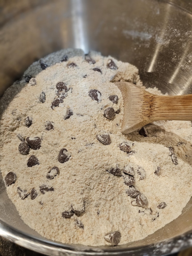 Puede incluir: Primer plano de un cuenco de acero inoxidable lleno de ingredientes para masa de galletas. La masa es de color marr&oacute;n claro, salpicada de chispas de chocolate negro. Una cuchara de madera descansa en la masa.