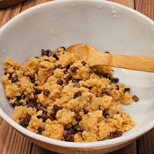 Puede incluir: Un cuenco de cer&aacute;mica blanco lleno de masa para galletas y chispas de chocolate. Una cuchara de madera descansa en la masa. El cuenco est&aacute; sobre una superficie de madera, con una tabla de cortar de madera y un electrodom&eacute;stico rojo en el fondo.