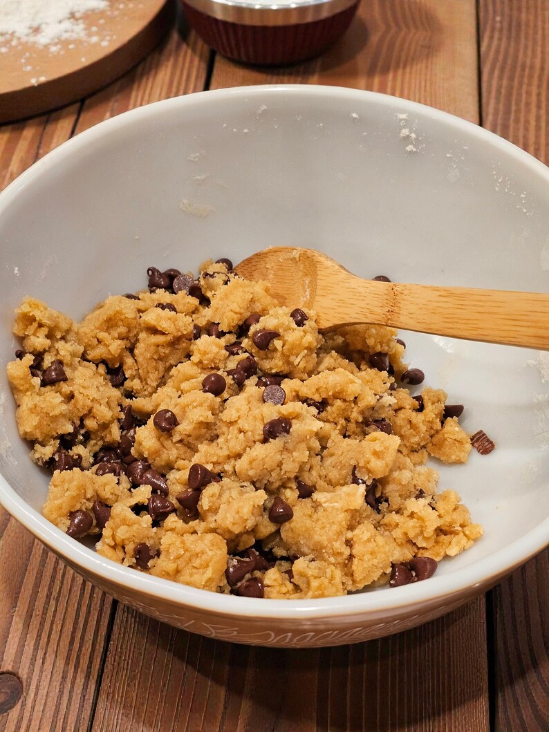 Puede incluir: Un cuenco de cer&aacute;mica blanco lleno de masa para galletas y chispas de chocolate. Una cuchara de madera descansa en la masa. El cuenco est&aacute; sobre una superficie de madera, con una tabla de cortar de madera y un electrodom&eacute;stico rojo en el fondo.