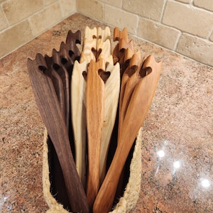 May include: A collection of wooden spatulas with heart-shaped cutouts in the handles, displayed in a woven basket. The spatulas vary in color, from dark brown to light tan. The basket is made of natural fibers and sits on a speckled countertop.