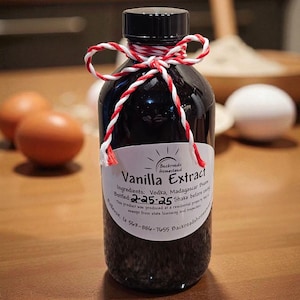 May include: A dark glass bottle of vanilla extract with a black cap, tied with red and white twine. The label reads "Vanilla Extract" and lists ingredients. The background includes eggs and baking ingredients.