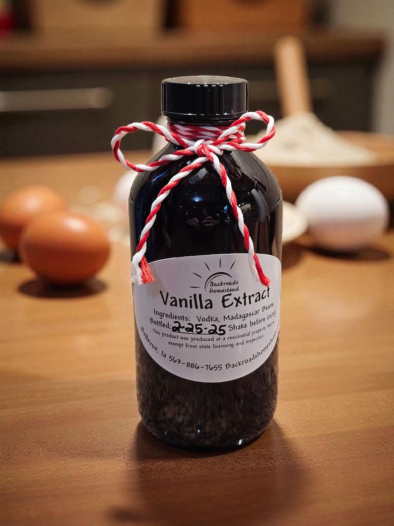 May include: A dark glass bottle of vanilla extract with a black cap, tied with red and white twine. The label reads "Vanilla Extract" and lists ingredients. The background includes eggs and baking ingredients.