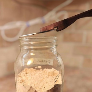 May include: A clear glass mason jar filled with a light beige powder, featuring a handwritten label that reads peanut butter. A dark brown wooden lid lifter with a metal hook is positioned at the top, partially lifting the metal lid from the jar.