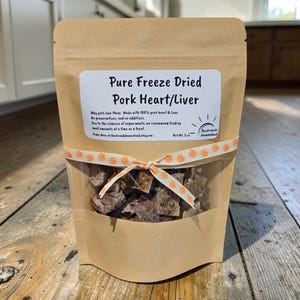 May include: A tan stand-up pouch containing freeze-dried pork heart and liver treats. The label reads Pure Freeze Dried Pork Heart/Liver, Net Wt. 2 oz, and includes a Backroads Homestead logo. A white ribbon with orange paw prints is tied around the bag.