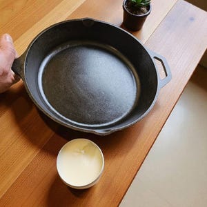 Puede incluir: Una sartén de hierro fundido negro con dos asas descansa sobre una superficie de madera. Un pequeño recipiente redondo con una sustancia blanca está al lado de la sartén. Una pequeña planta suculenta en maceta también está sobre la superficie de madera.