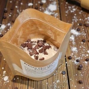 Puede incluir: Una bolsa de papel marr&oacute;n llena de mezcla para galletas de chocolate artesanal, salpicada de chispas de chocolate y harina sobre una superficie de madera. Un rodillo de madera est&aacute; en el fondo. La etiqueta de la bolsa es parcialmente visible.