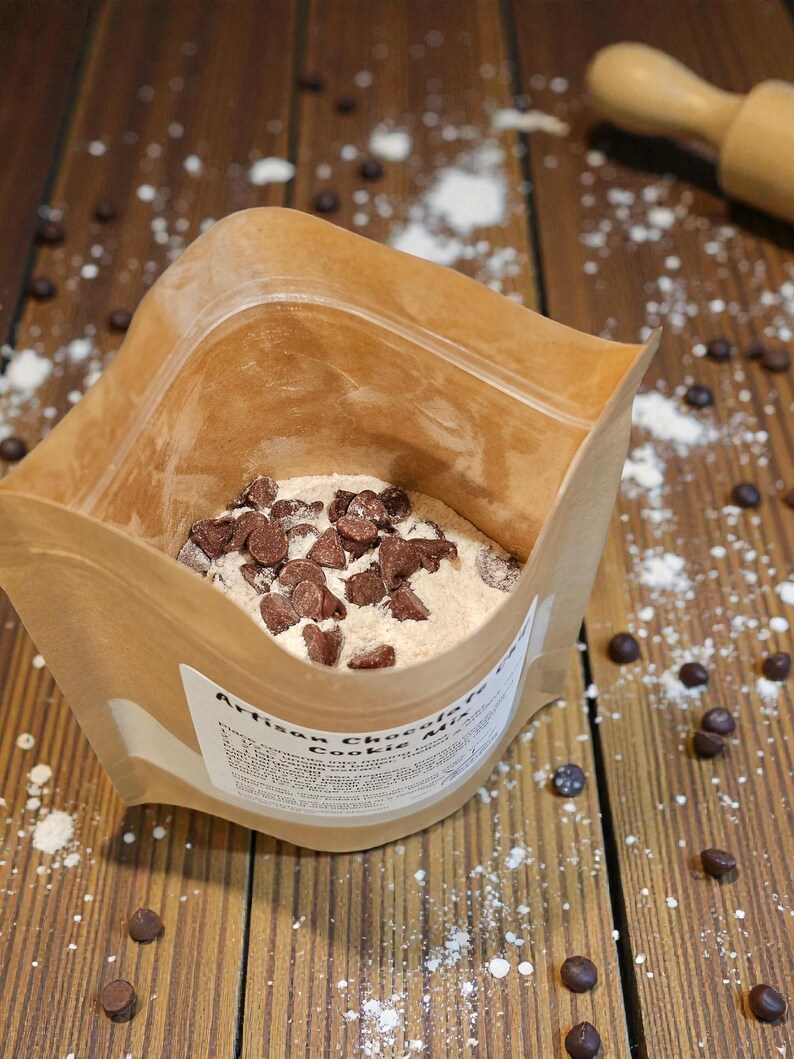 Puede incluir: Una bolsa de papel marr&oacute;n llena de mezcla para galletas de chocolate artesanal, salpicada de chispas de chocolate y harina sobre una superficie de madera. Un rodillo de madera est&aacute; en el fondo. La etiqueta de la bolsa es parcialmente visible.