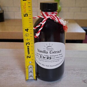 May include: A dark glass bottle of vanilla extract, approximately 5 inches tall, with a black cap and a red and white twine tie. The label reads "Vanilla Extract" and lists ingredients and bottling date.
