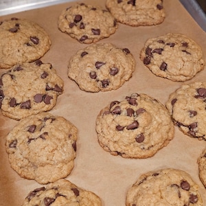Puede incluir: Una bandeja para hornear con una docena de galletas con chispas de chocolate reci&eacute;n horneadas. Las galletas son de color marr&oacute;n dorado con chispas de chocolate visibles. Est&aacute;n dispuestas sobre papel de hornear.