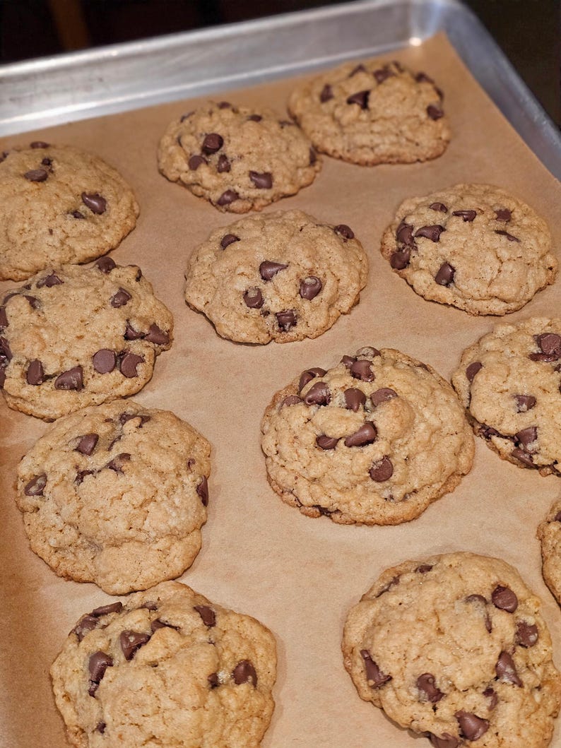 Puede incluir: Una bandeja para hornear con una docena de galletas con chispas de chocolate reci&eacute;n horneadas. Las galletas son de color marr&oacute;n dorado con chispas de chocolate visibles. Est&aacute;n dispuestas sobre papel de hornear.