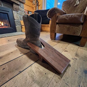 Puede incluir: Una bota de trabajo de cuero marrón se coloca en un sacabotas de madera. El sacabotas es de madera oscura y tiene una ranura para el talón. La bota está sobre un suelo de madera.