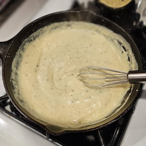 Puede incluir: Una sartén de hierro fundido llena de salsa cremosa y clara, posiblemente bechamel o similar. Un batidor de acero inoxidable está parcialmente sumergido en la salsa, lo que sugiere que se está removiendo. La sartén está sobre una estufa negra.
