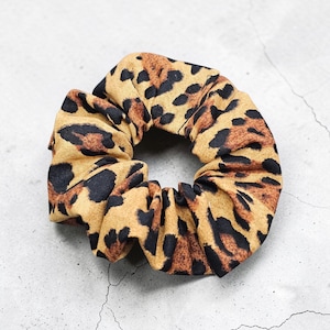 May include: A leopard print scrunchie. The hair accessory features a tan fabric base with dark brown and black spots. The scrunchie is gathered and circular, designed for hair styling.