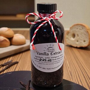 May include: A dark glass bottle of vanilla extract with a black cap, tied with red and white twine. The label reads "Vanilla Extract" and lists ingredients. Vanilla beans and coffee beans are on a black surface.