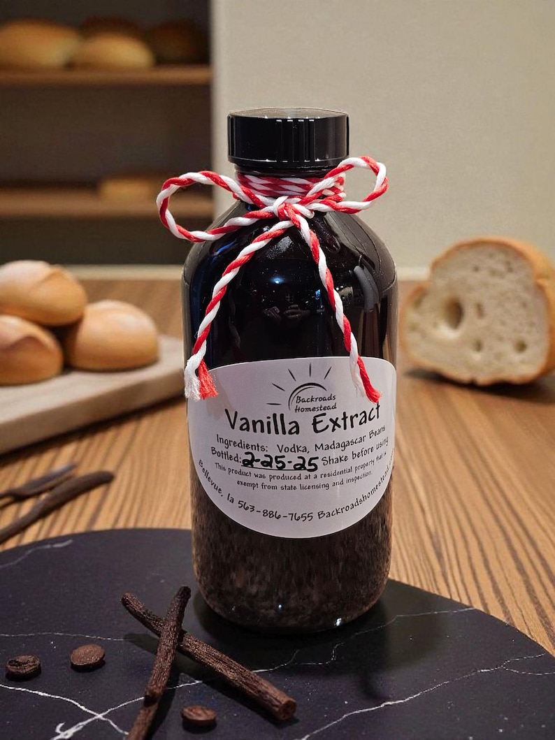 May include: A dark glass bottle of vanilla extract with a black cap, tied with red and white twine. The label reads "Vanilla Extract" and lists ingredients. Vanilla beans and coffee beans are on a black surface.