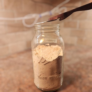 Op de afbeelding: Een heldere glazen pot gevuld met lichtbruin poeder, met de hand beschreven als "beauty". Een houten en metalen gereedschap bevindt zich boven de pot, wat duidt op het meten of doseren van de inhoud. De pot staat op een gespikkelde ondergrond.