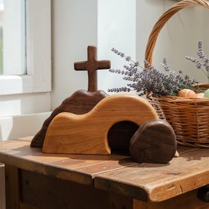 Può includere: Scena pasquale in legno con una croce in cima a una collina, un sepolcro con un ingresso aperto e una pietra. Un cestino di vimini con lavanda e uova di Pasqua è posto vicino su un tavolo di legno. La scena è illuminata dalla luce naturale.