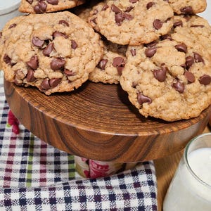 Puede incluir: Una pila de galletas con chispas de chocolate reci&eacute;n horneadas sobre un soporte de madera para pasteles. Las galletas son de color marr&oacute;n dorado con chispas visibles. Un vaso de leche est&aacute; al lado, junto con un pa&ntilde;o a cuadros.