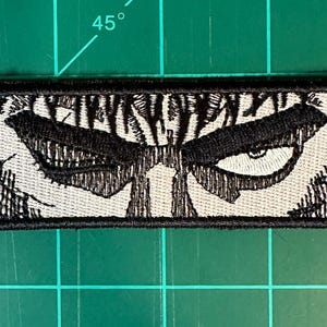 May include: A rectangular embroidered patch featuring a black and white graphic illustration of a pair of intense, narrowed eyes with heavy brows and detailed shading, framed by a thick black stitched border on a white fabric background.