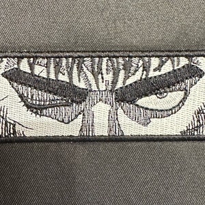 May include: Black and white embroidered patch featuring a close-up of an eye with a menacing expression.