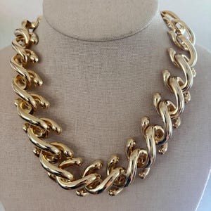 Puede incluir: Un collar de cadena en tono dorado grueso con eslabones grandes, pulidos y entrelazados. El collar tiene una apariencia pesada y redondeada con un acabado metálico de alto brillo, creando un estilo audaz que descansa en forma curva.