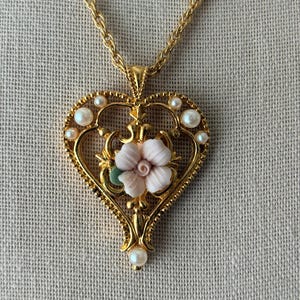 May include: A gold-tone heart-shaped pendant necklace featuring a central pale pink carved flower with a small green leaf. The heart frame is decorated with small white faux pearls and intricate metal scrollwork, suspended from a gold chain.