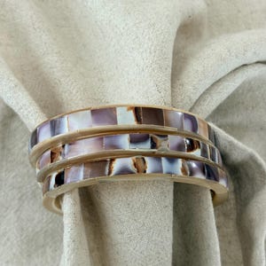 Op de afbeelding: Een set van drie ronde servetringen met een mozaïekinleg van rechthoekige stukjes in bruin-, crème- en iriserende paarstinten. De ringen zijn voorzien van een gladde, lichtgekleurde rand die de patronen in het midden van de ringen omlijst.