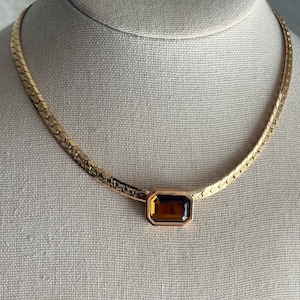 May include: A gold-toned herringbone chain necklace featuring a central rectangular pendant with a faceted amber-coloured stone. The chain has a textured, woven appearance, and the pendant is set in a smooth, polished gold-toned metal frame.