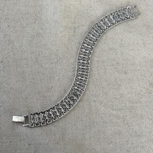 Op de afbeelding: Een zilverkleurige metalen schakelarmband met een herhalend geometrisch patroon van onderling verbonden rechthoekige segmenten. De armband heeft een gepolijste afwerking en is voorzien van een veilige rechthoekige baksluiting aan één uiteinde.