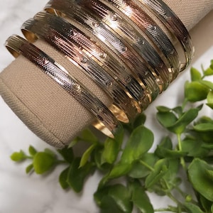 Op de afbeelding: Een stapel gouden, zilveren en roségouden armbanden met een gegraveerd ontwerp. De armbanden zijn gestapeld op een beige oppervlak.