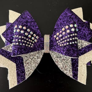 May include: Purple and silver cheer bow with rhinestones. The bow is made of glitter fabric and has a silver center.