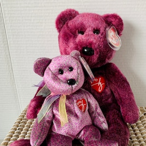 May include: Two purple Beanie Babies, one with a floral pattern and the other with a solid colour, are sitting together. The floral Beanie Baby has a yellow ribbon around its neck and a red heart tag with the year 2000 on it. The solid purple Beanie Baby has a red heart tag with the Ty logo on it.
