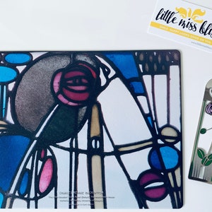 Op de afbeelding: Een kleurrijk glas-in-loodontwerp met geometrische vormen en abstracte patronen in tinten blauw, rood en bruin. Het ontwerp is omlijst door een zwarte rand en bevat de tekst "Charles Rennie Mackintosh".