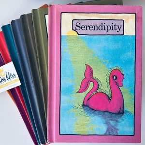 May include: A stack of ten colourful hardback books with the title "Serendipity" on the top book. The book cover features a pink sea serpent with green seaweed on its back swimming in blue water.