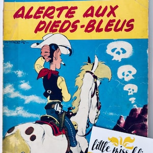 May include: A vintage Lucky Luke comic book, number 10, titled "Alerte aux Pieds-Bleus." The cover shows a cowboy on a white horse against a blue sky with skull graphics. The title is in yellow and red, and the text "little miss bliss" is also visible.