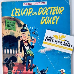 May include: A vintage comic book cover for Lucky Luke VII, titled "L'Elixir du Docteur Doxey." The cover is predominantly blue with yellow text and illustrations of cowboys and a doctor. A business card with the text "little miss bliss" is also visible.