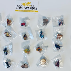 Puede incluir: Una colección de 20 pines de esmalte coloridos en bolsas de plástico transparente individuales. Los pines presentan varios diseños de animales y naturaleza, incluyendo una mariposa, un pez, una tortuga, una araña, un pájaro, un mono y un oso. Los pines están dispuestos en cuatro filas de cinco, con el texto "little miss bliss" visible en la parte superior de la imagen.