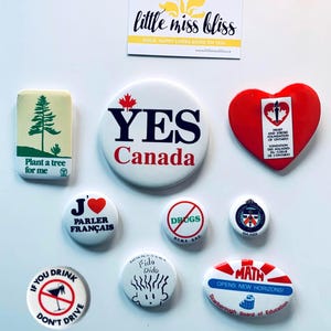 May include: A selection of badges and a small rectangular magnet. The badges display diverse designs, including text such as "YES Canada," "J'❤ PARLER FRANÇAIS," and "MATH OPENS NEW HORIZONS!" The magnet reads "little miss bliss."