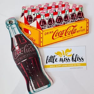 Puede incluir: Una caja vintage de Coca-Cola con 24 botellas de Coca-Cola. La caja es amarilla con detalles rojos y blancos. El texto en la caja dice "Drink Coca-Cola in Bottles". Hay una gran botella de Coca-Cola con una etiqueta roja y blanca.