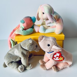 May include: Four colourful plush toys, including a rainbow bear, a rainbow bunny, a brown bear and a pink pig, are arranged on a yellow box. The pig has a red and white Ty tag attached to it.