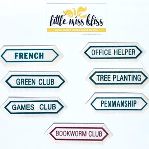 Könnte beinhalten: Eine Sammlung weißer Stoffetiketten mit teal- und burgunderfarbenen Rändern, jeweils mit einem anderen Text. Die Texte lauten "FRENCH", "OFFICE HELPER", "GREEN CLUB", "TREE PLANTING", "GAMES CLUB", "PENMANSHIP" und "BOOKWORM CLUB". Ein Logo ist oben.