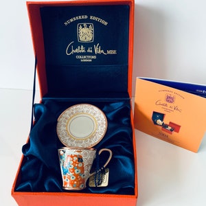 Puede incluir: Una caja roja con un interior de satén azul que contiene una taza de té y un platillo blancos y dorados con un diseño floral colorido. La caja tiene una etiqueta dorada que dice "Charlotte di Vita Collections London MBE Numbered Edition".