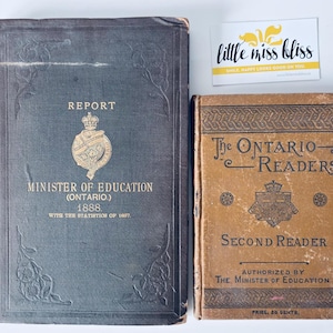 Puede incluir: Se muestran dos libros antiguos. El libro más grande es azul oscuro con letras doradas que dicen "REPORT" y "MINISTER OF EDUCATION (ONTARIO.) 1888." El libro más pequeño es de color marrón claro y se titula "The ONTARIO READERS SECOND READER." También hay una pequeña tarjeta con el texto "little miss bliss".