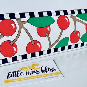 May include: Black and white striped border with red cherries and green leaves on a white background. The border is decorated with a repeating pattern of cherries and leaves. The text "little miss bliss" is printed on a yellow background with the tagline "smile, happy looks good on you." and the website address "www.littlemissbliss.ca".