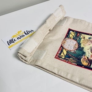 May include: A beige canvas bag with a red, green, and yellow print featuring a girl and the text "UNTO OTHERS AS YOU WOULD HAVE THEM DO UNTO YOU". The bag has white velcro closures.