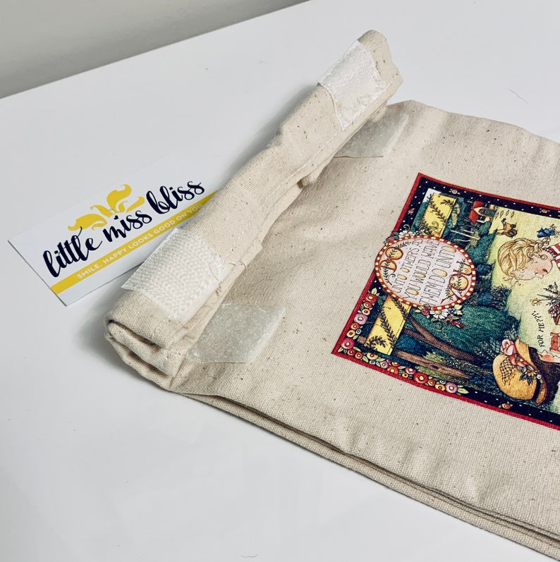 May include: A beige canvas bag with a red, green, and yellow print featuring a girl and the text "UNTO OTHERS AS YOU WOULD HAVE THEM DO UNTO YOU". The bag has white velcro closures.