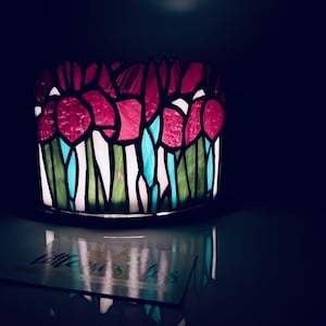 May include: A stained glass candle holder with a floral design. The design features red, white, green, and blue glass pieces. The candle holder is illuminated from within, creating a soft glow. The overall theme is home decor.