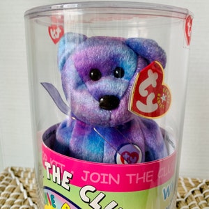 Può includere: Un orsacchiotto Beanie Baby tie-dye blu e viola in un contenitore di plastica trasparente con un nastro viola intorno al collo. L'orso ha un'etichetta Ty rossa attaccata all'orecchio e un'etichetta Ty viola sul petto. Il contenitore ha un'etichetta rosa e bianca che dice "Join the Club".