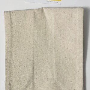 May include: A beige canvas fabric with a speckled texture. The fabric is folded in half and appears to be a piece of material for crafting or sewing projects.