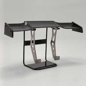 Peut inclure: Un aileron arrière en fibre de carbone noire avec deux jambes de force réglables en métal argenté. L'aileron est monté sur un support en métal noir avec le texte "3Racing" imprimé sur le devant.